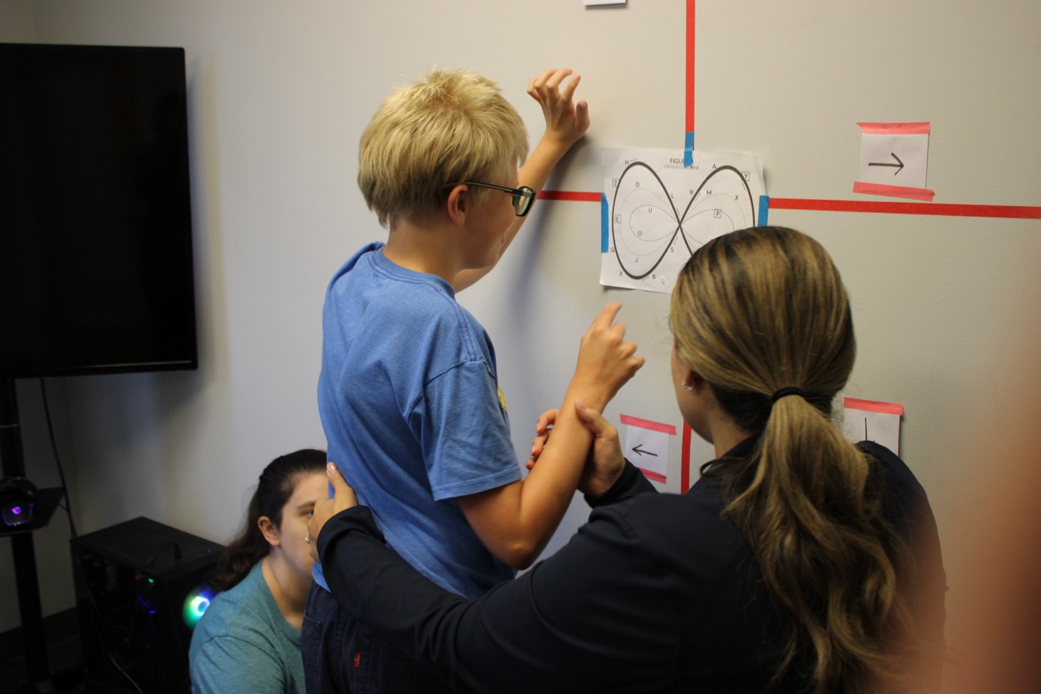 A pediatric therapist guides a child's arm through a figure-eight infinity midline-crossing exercise on a wall chart during a session at Bright Beginnings in Marlton, NJ